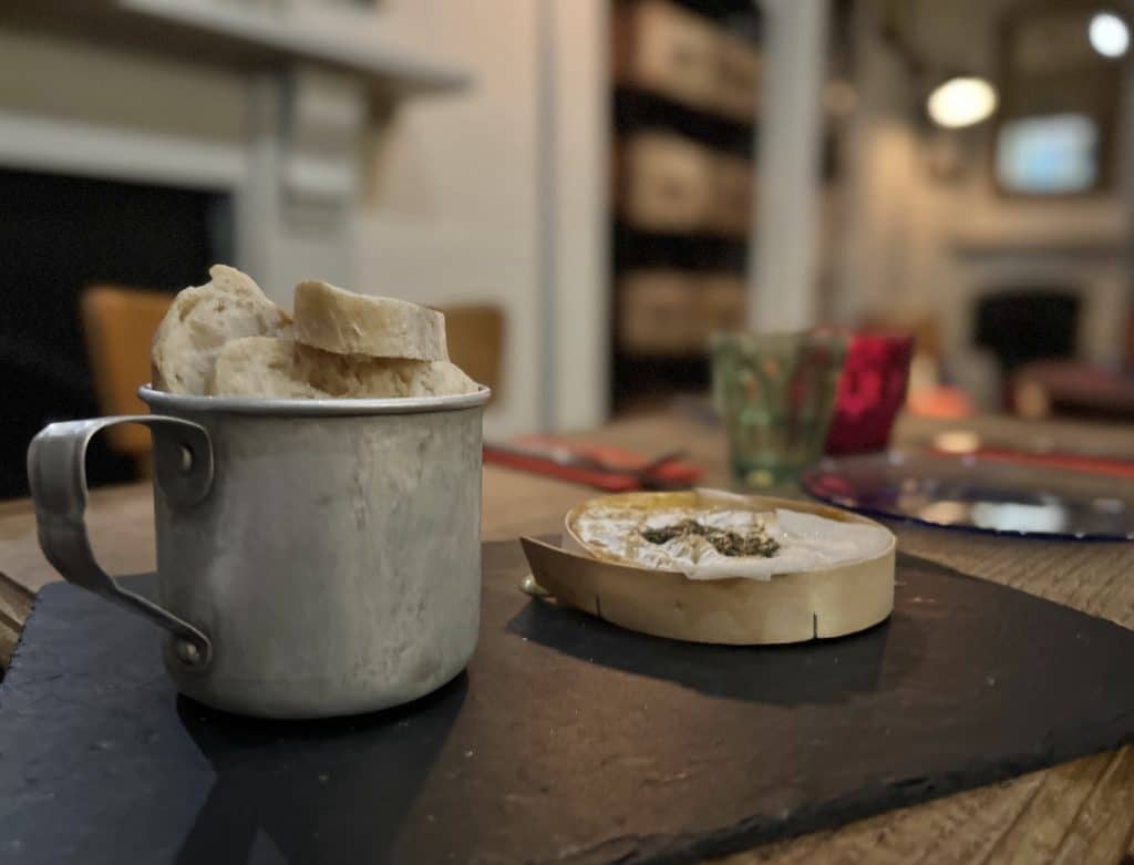 Camembert at Comptoir + Cuisine in Bath. A small Camembert wrapped in wood and a silver mug full of warm b read on a dark slate with red and pale green glasses in the background.
