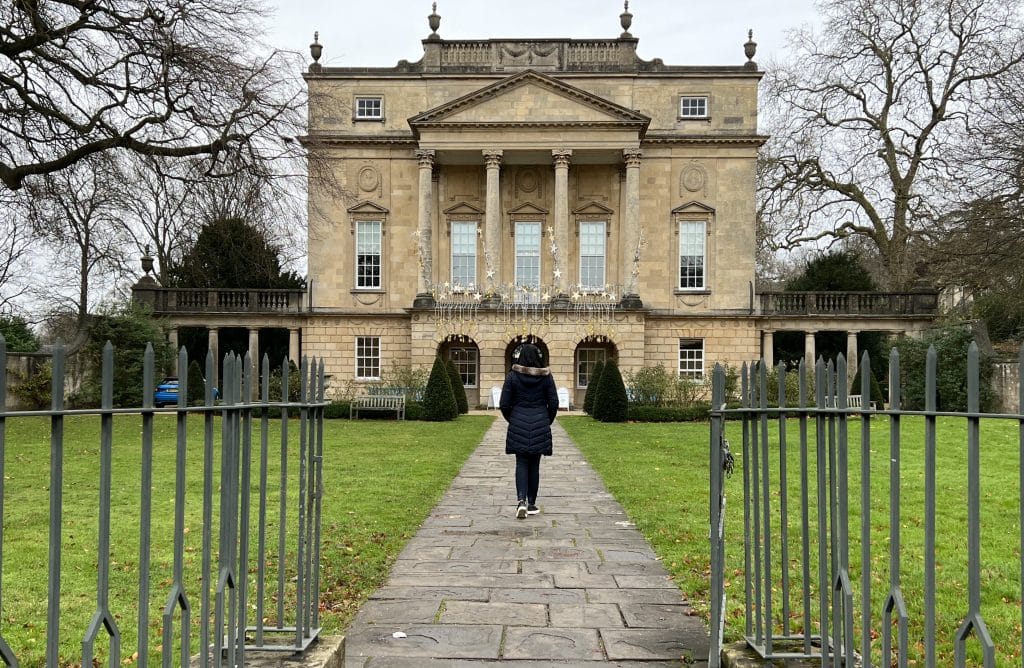 Bejal walking down the from aisle of the Holburne Museum, Bath. There are bare trees at either end in December and Bejal is wearing a blue winter coat