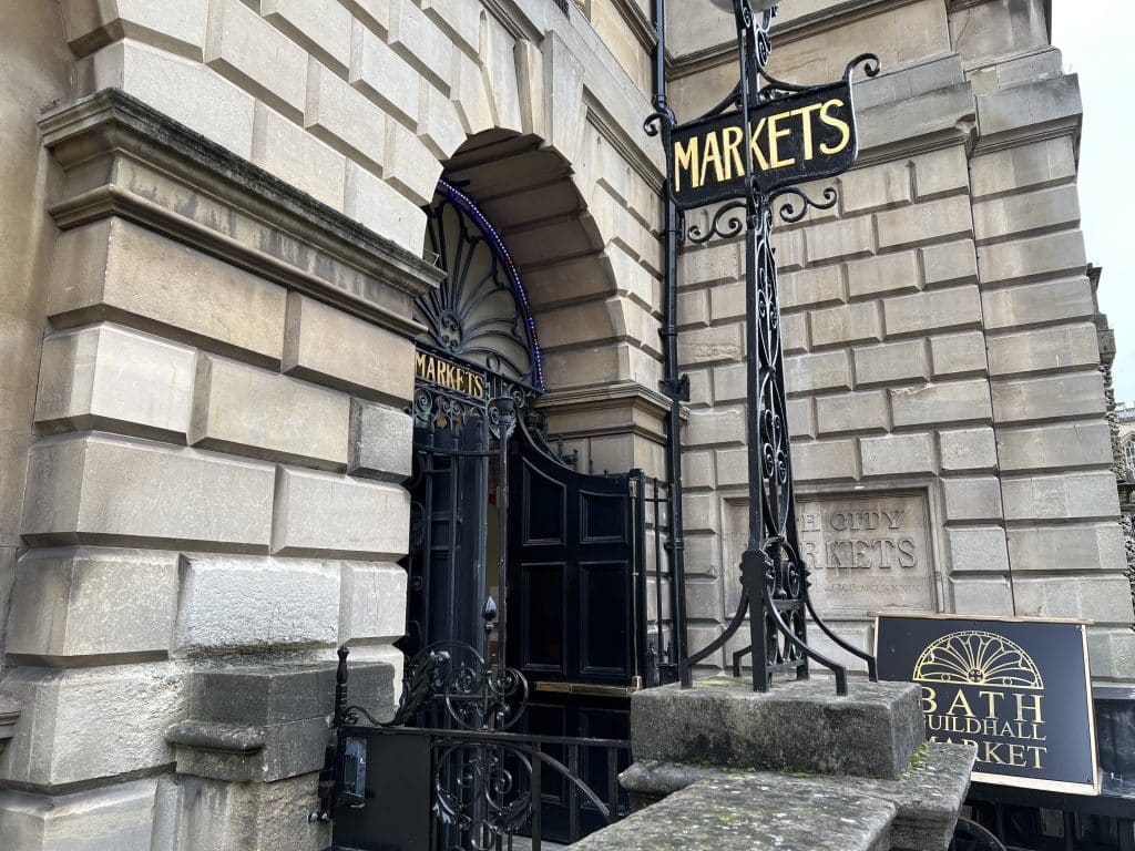 The exterior of the Guildhall Market, Bath with black signs and gold lettering reading 'markets'.