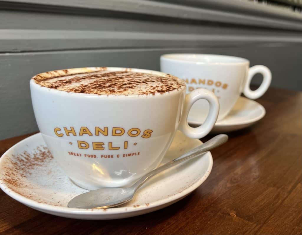 Coffees at Chandos, Bath. Two white cups of coffee with saucers containing cappuccino dusted with chocolate on a wooden table. One of teh saucers has a tea spoon. 
