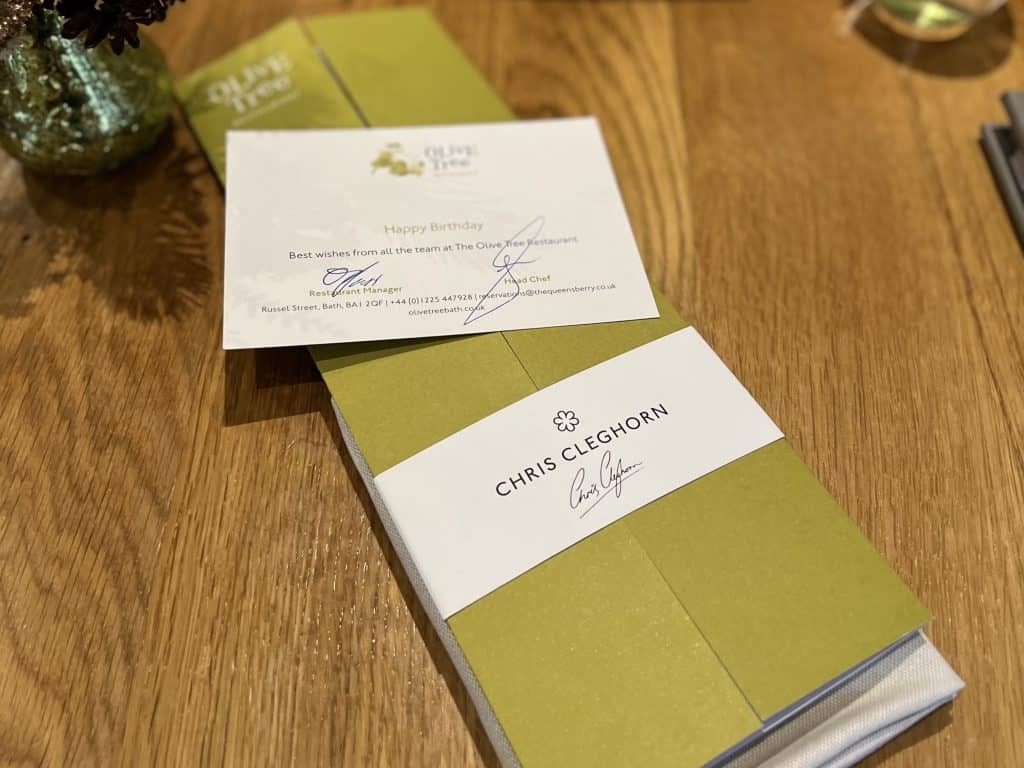 Olive Tree, Bath Menu signed by Chris Cleghorn in a sage and light cream colour placed on a light wood grain table at Queens berry Hotel, Bath