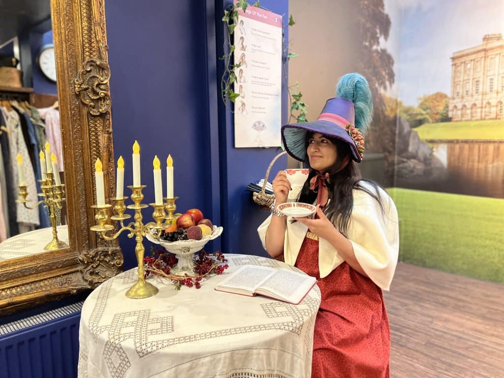 The Jane Austen Centre in Bath. Bejal is wearing a red regency outfit with a blue bonnet sipping on a tea at the Jane Austen Centre. The table has a fake fruit bowl with a gold candelabra with candles.
