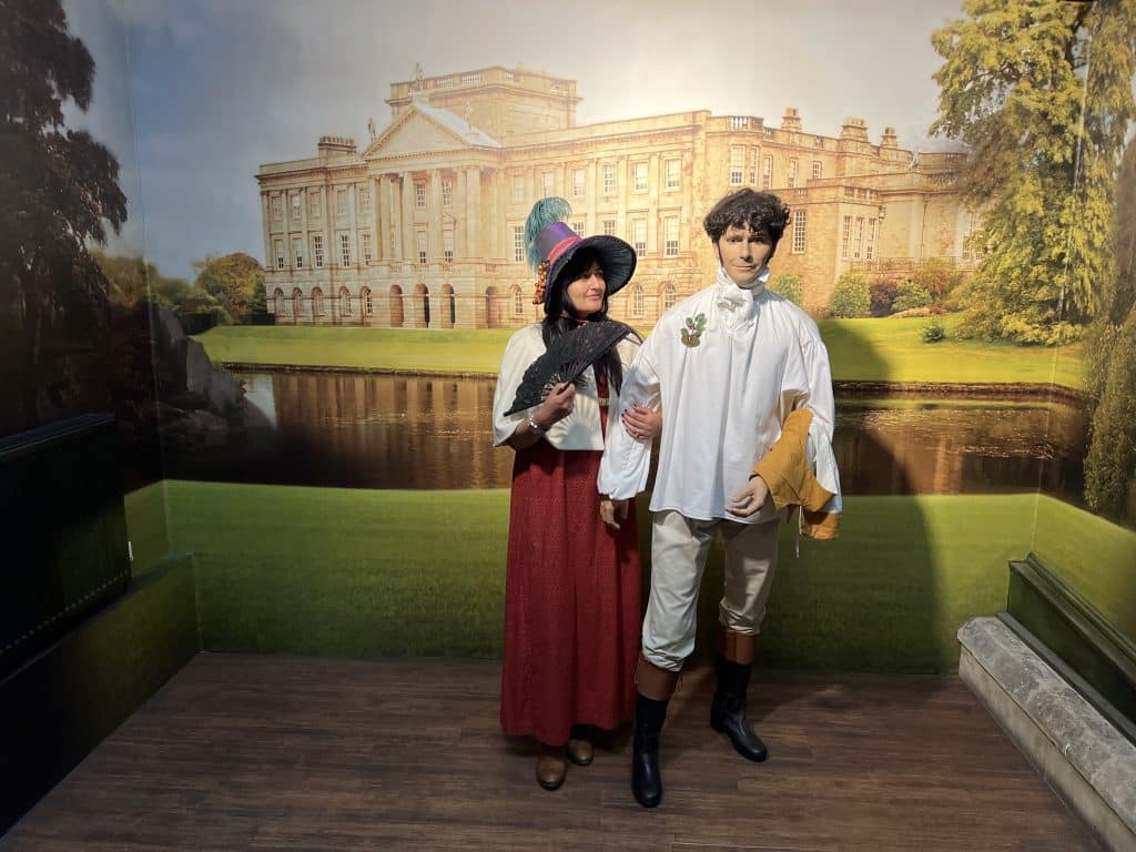 Bejal wearing a red Regency dress, white cape and blue and pink bonnet  next to a Mr Darcy mannequin with a Regency-style background house and gardens at The Jane Austen Centre, Bath