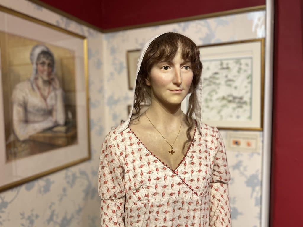 A wax work of Jane Austen wearing a cream dress with maroon florals against a red wall. She is wearing a veil over her brown hair and wears a gold cross necklace, at the Jane Austen Centre, Bath