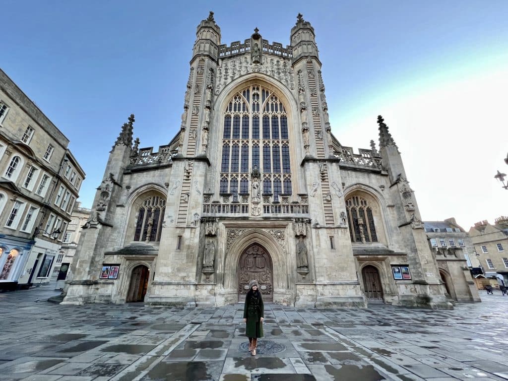 Bejal standing outside Bath Abbey on damp paving stones wearing a faux fur hat, khaki winter wool coat and tan boots. The sky is blue in the background