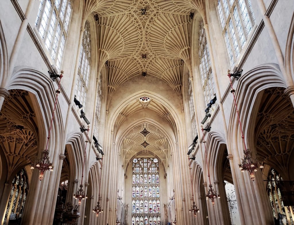 The interior ceiling of Bath Abbey with its white intricately decorated interiors and light coming in through the stained glass window behind the alter.
