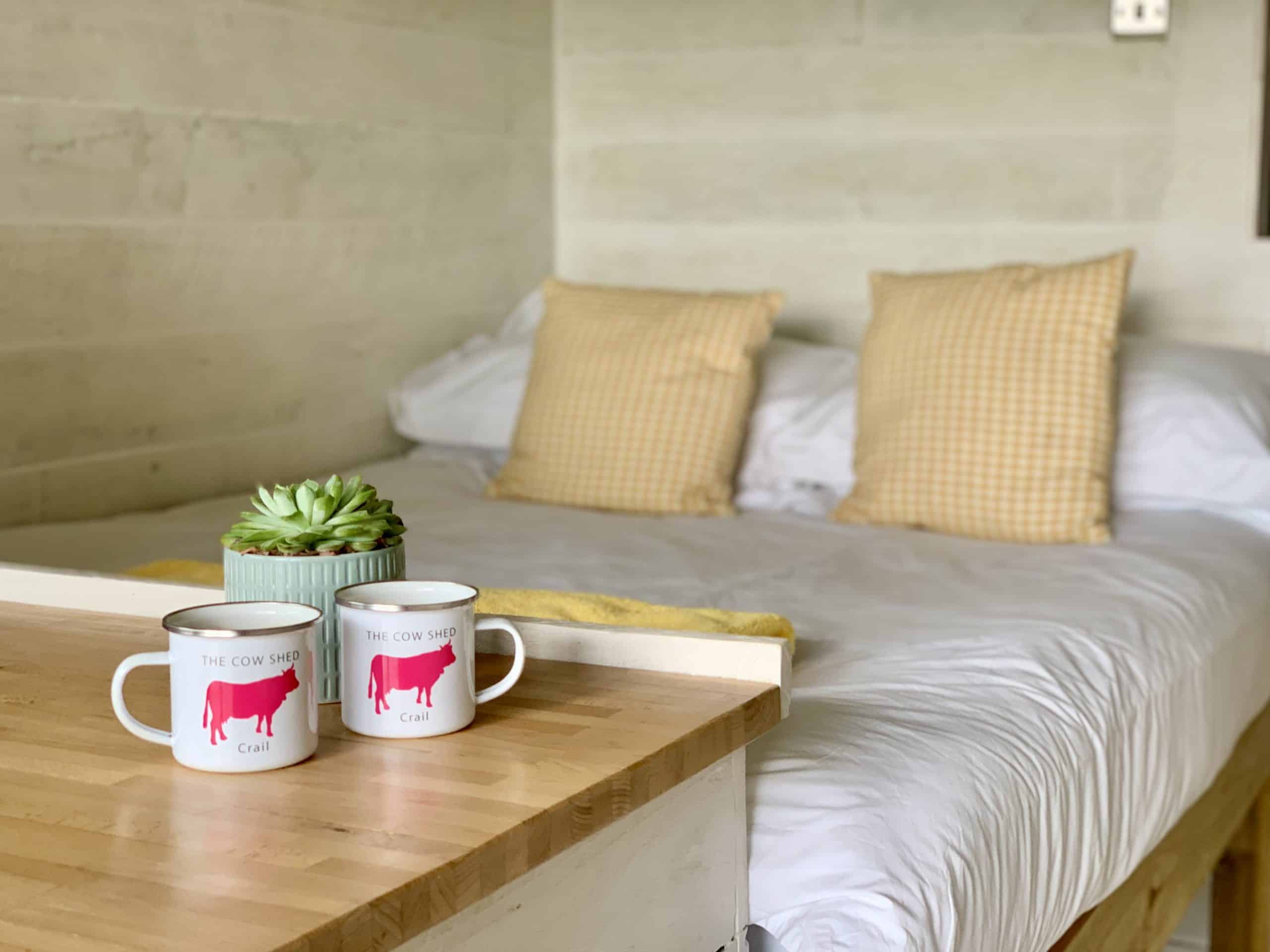 Cow Pad Daisy, bed, Crail with a plant and 2 mugs of tea on a worktop. The bedding is crisp white with yellow checked cushions.