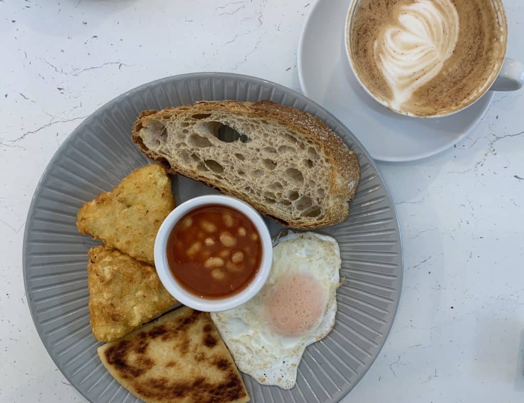 Em's Kitchen top lay of a vegetarian breakfast with a fried egg, slice of bread, beans, hash browns and a cappuccino. Located in Edinburgh