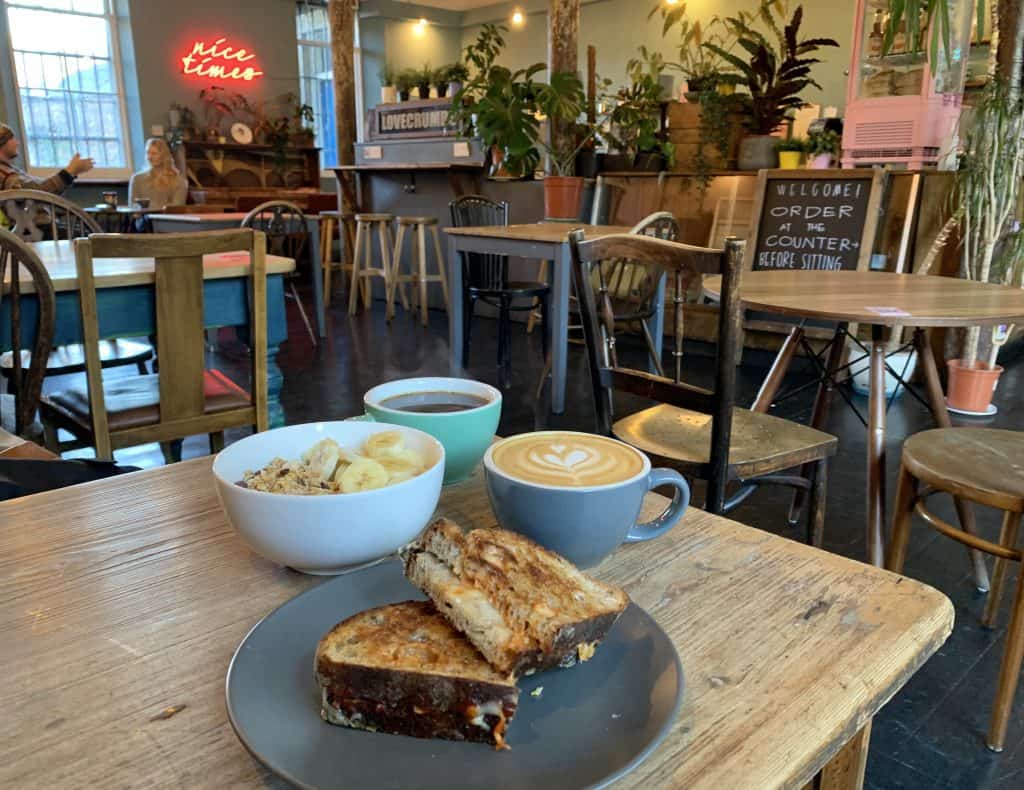Lovecrumbs Cafe with a bowl of granola, 2 coffees and 2 slices of banana bread. The cafe has quirky furniture in the background and a neo 'nice times' sign Located in Edinburgh