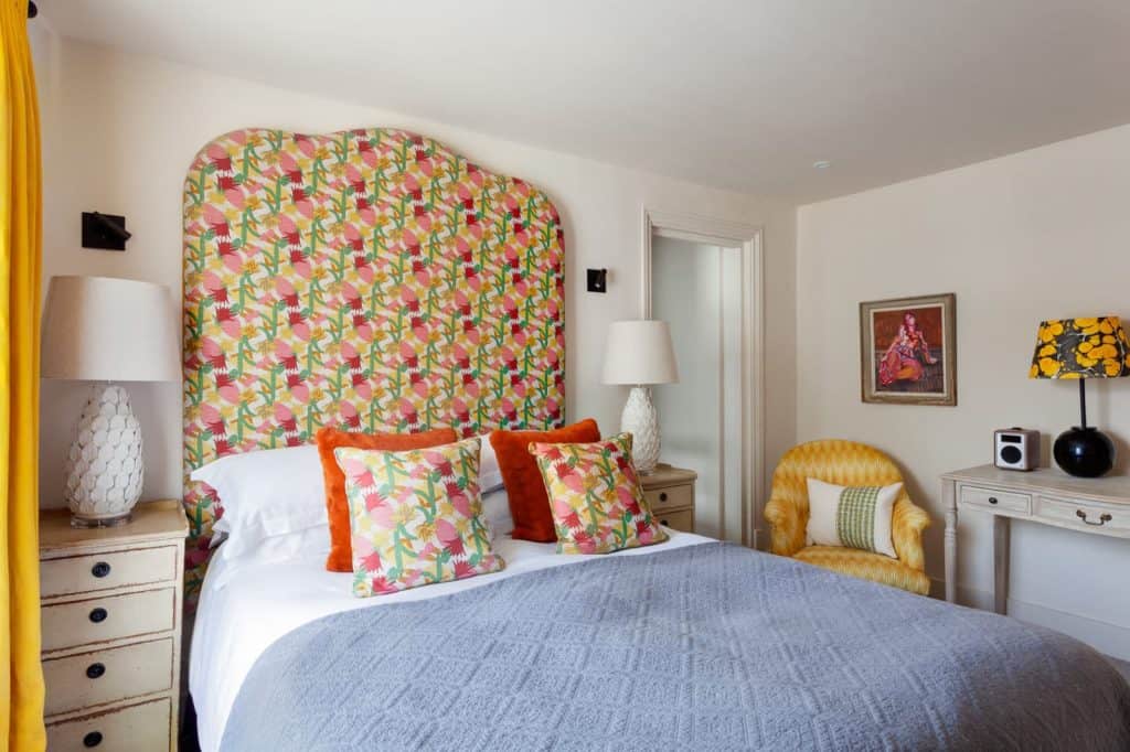 Classic Double Room at The Falstaff, Canterbury. The headboard is green, cerise pink and yellow to match the scatter cushions on the bed which has white linen and a pale blue throw. There is a white side table and a light orange arm chair to the side