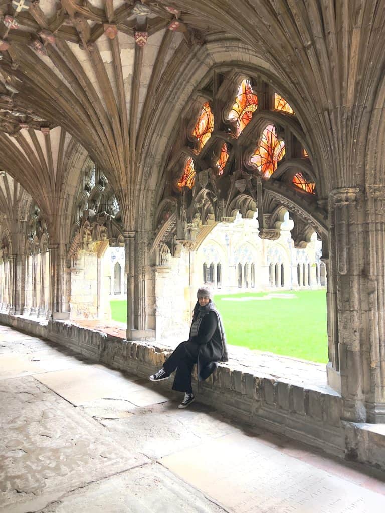 Bejal sitting in Canterbury cathedral Cloisters with stained glass windows and grassy courtyard in the background