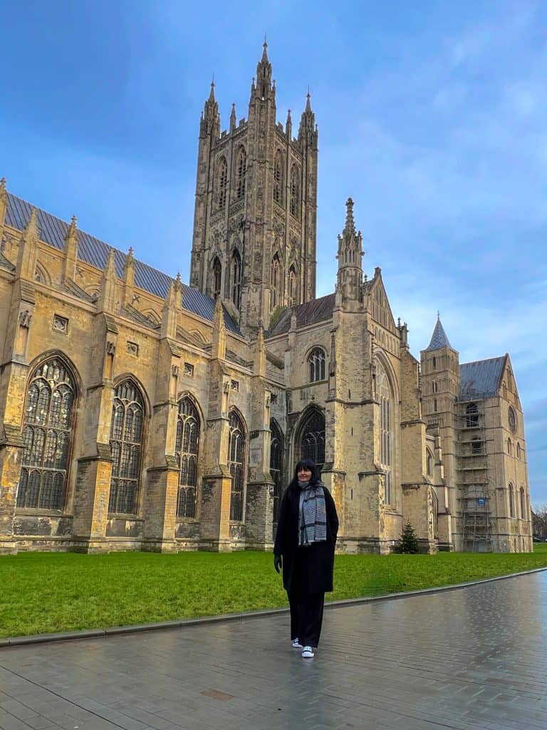 Front of Canterbury Cathedral Exterior with Bejal standing outside wearing a black coat with checked black and white scarf