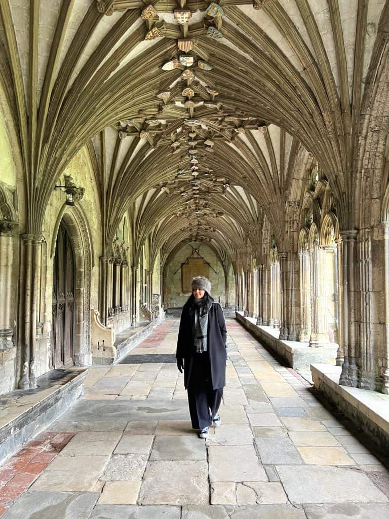 Bejal walking through Cathedral Cloisters quarters in Canterbury wearing a faux fur hat and a black coat with black and white oversized scarf