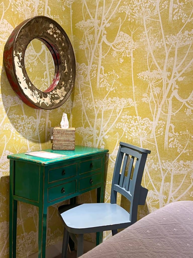 The Falstaff, Canterbury: Cosy Room  with yellow and white wallpaper, wooden. beams. A teal table stands at the side with a grey chair and a bronze framed circular mirror.