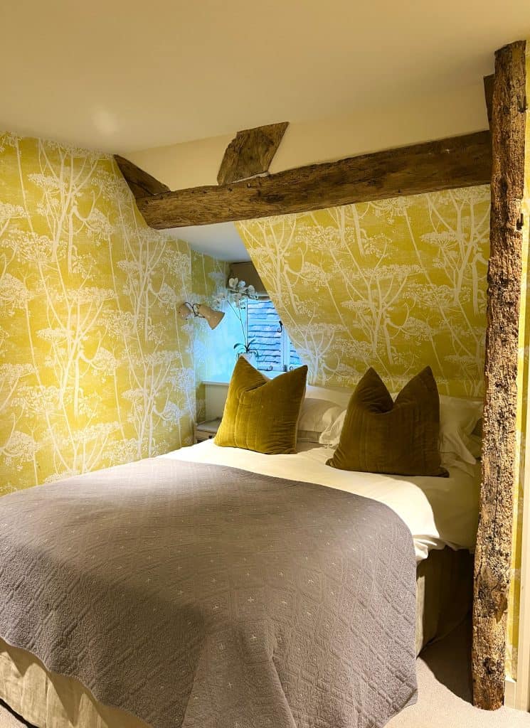 The Falstaff, Canterbury: Cosy Room  with yellow and white wallpaper, wooden. beams, a grey throw on the bed and a taupe coloured scatter cushions. The bed linen is white.