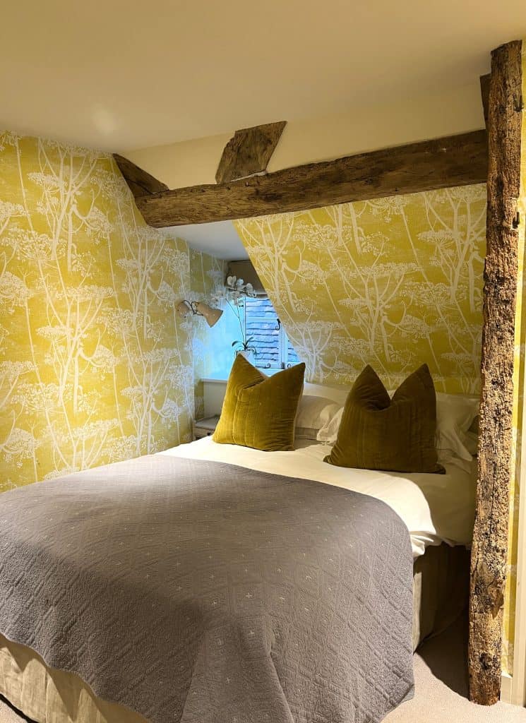 The Falstaff cosy room with bed and scatter cushions, beamed ceilings and mustard walls featuring white plant stem prints.