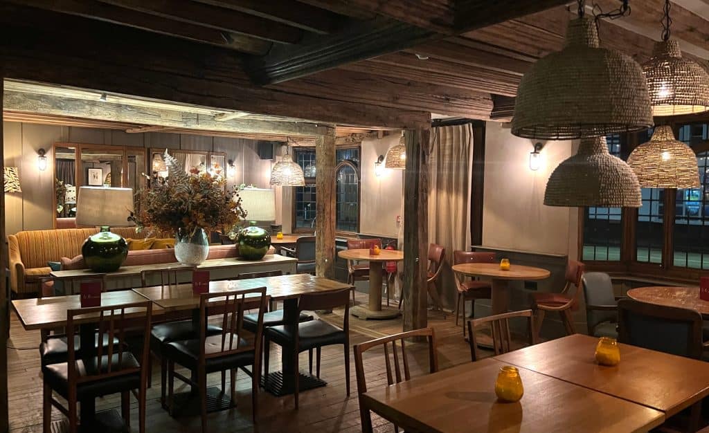 The Falstaff, Canterbury: dining/lounge rooms with tables and sofas in different tones of wood with rattan pendant lights. There are mustard coloured sofas in the background