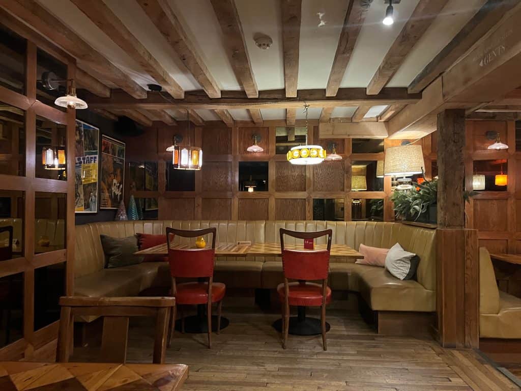 The Falstaff, Canterbury dining room with diner style seating with beige upholstered cushioned booths and red upholstered high back dining chairs. There is low level pendent lighting and low wooden ceilings