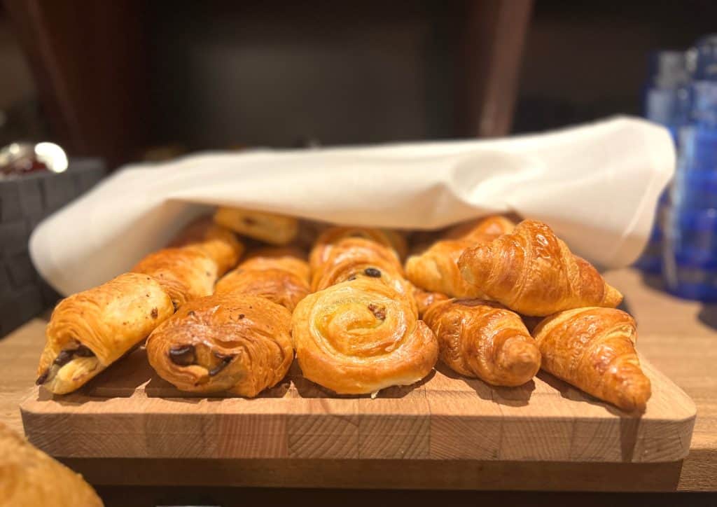 The Falstaff, Canterbury: Breakfast Pastries, pain au chocolat, danish pastries and croissants. They are on a wooden board and half covered with a white linen napkin.