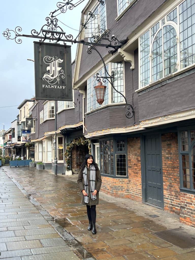 Bejal standing outside the entrance of The Falstaff, Canterbury on a rainy day wearing a duffle coat and black and white checked scarf