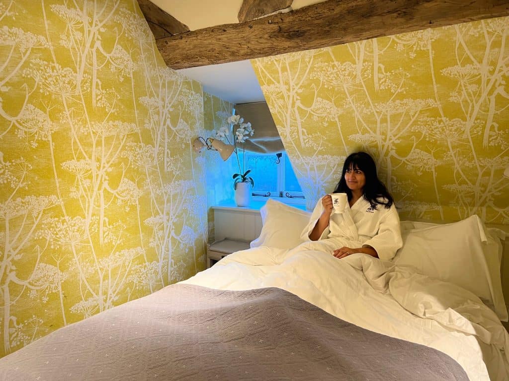 Bejal in white robe in the cosy room bed with a coffee mug in hand. The walls are mustard and white with wooden beams above here and there is a white orchid on the window ledge. The bed linens are white with a grey throw half draped at the end of the bed.