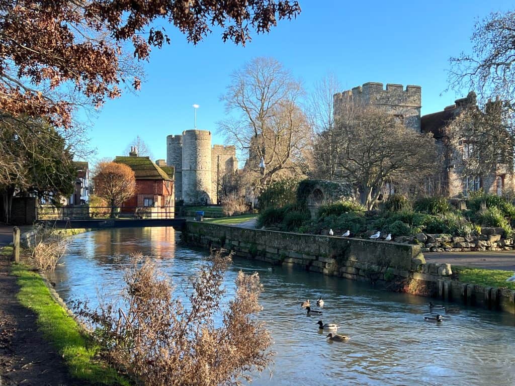 Westgate gardens with Westgate Towers and the River Stour. There are ducks in the river and dry foliage on the banks of the river.