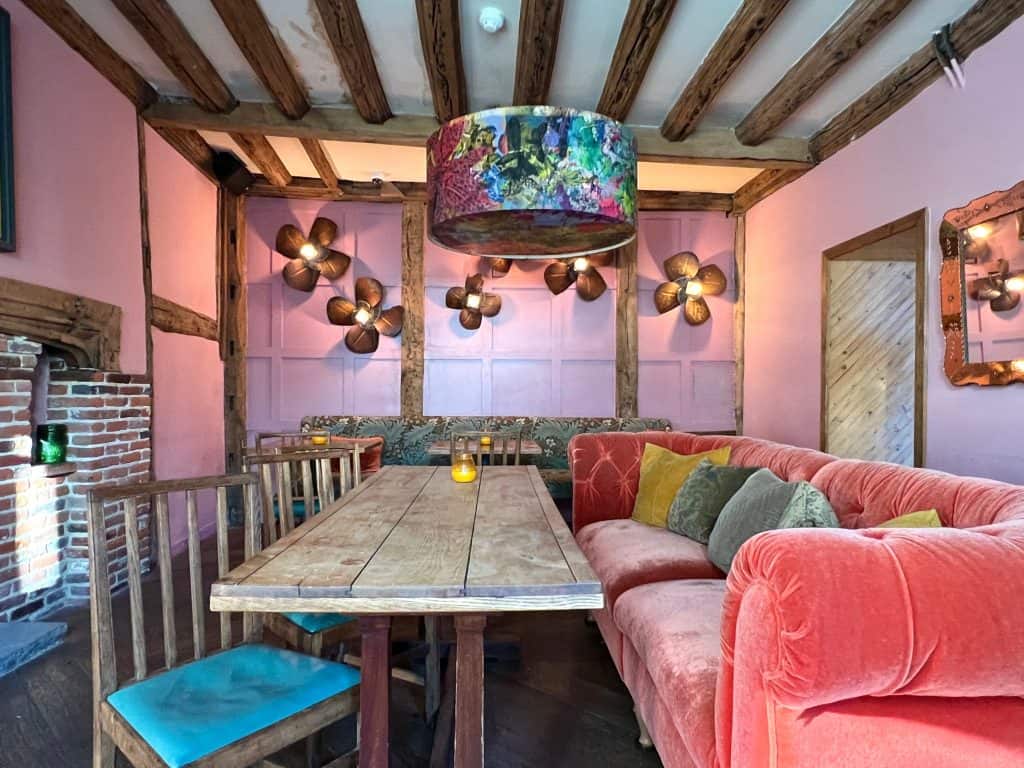 Drawing room with pink and teal sofas and chairs and a wooden table in the centre with teal cushions on the chairs at The Falstaff, Canterbury. There is a pink, teal and lavender light shade hanging from the wooden low beam ceiling. The walls are a pale pink colour