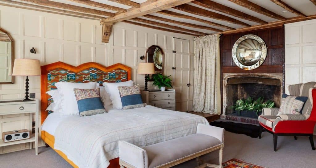 Junior Suite at The Falstaff, Canterbury, The bed in made with white linen and orange, red and blue scatter cusions which match teh headboard. There is a red striped chair near the fire place and a bedtide table with a potted plant. 