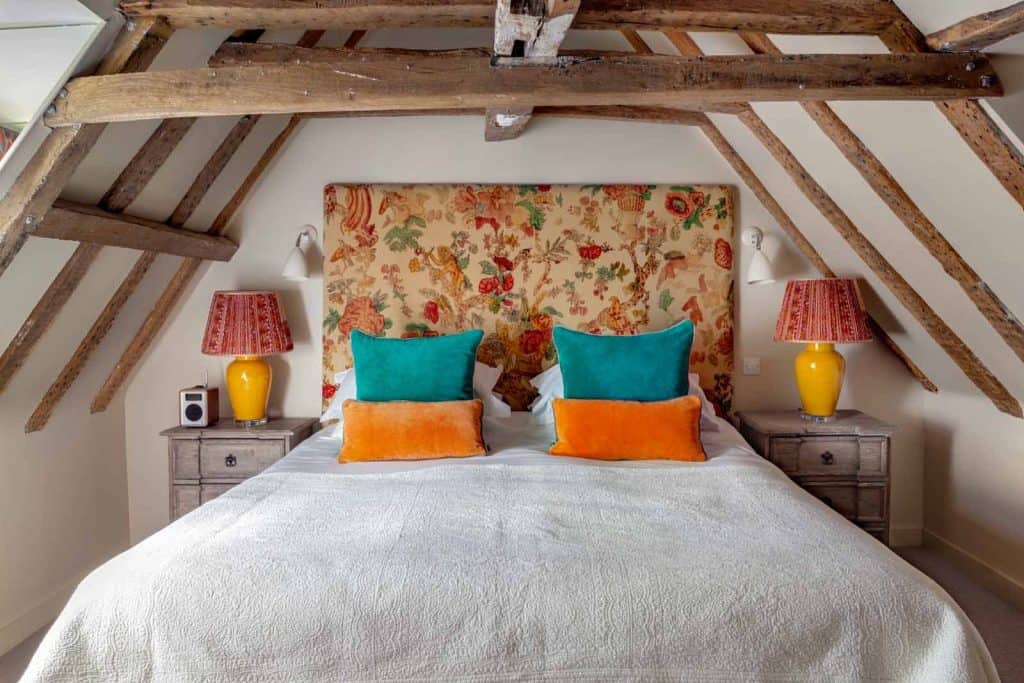 Luxury Double at The Falstaff, Canterbury. There is a wooden beamed ceiling with white walls. The bed has white linens and teal and oranhe scatter cusions. The two bedside tables have pink lampshades and yellow lamp bases with a floral upholstered headboard in complementing colours.