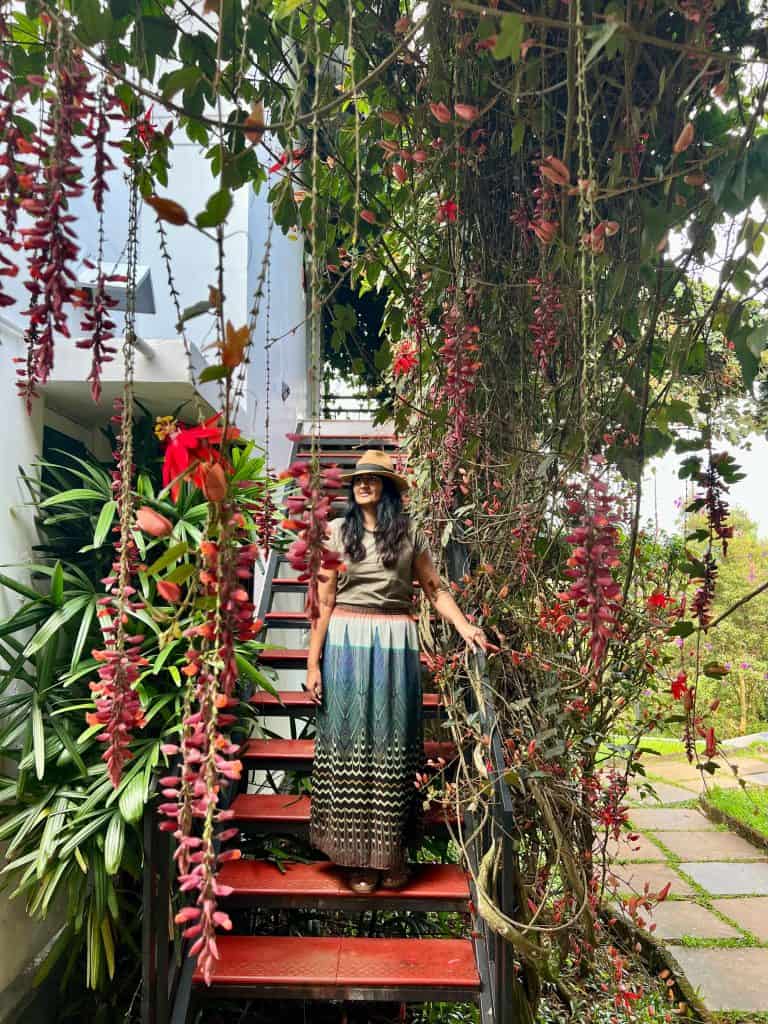 Eco-friendly Hotels in Kerala: Bejal standing on stairs pf a cottage with the read leaved plant draping from the tree above her. beja; is wearing a khaki T-shirt with a green, purple and orange skirt and a tan coloued hat.