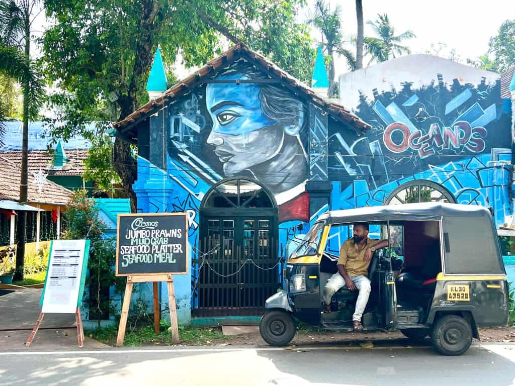 Man in his rickshaw outside the street art covered Oceanos restaurant. He is wearing white jeans and a pale gold shirt.