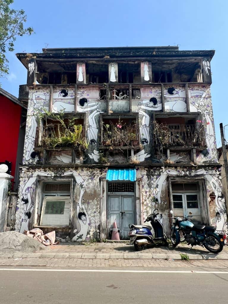 A three story old distressed building in Fort Kochi with overgrown plants on the windows, mismatched exteriors and a motorbike outside it.