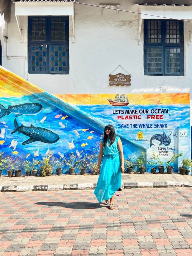 Kerala one of India's most eco-friendly states: Bejal standing in front of eco-friendly sign in Fort Kochi. The wall behind has ocean street art with dolphins and marin dlife. It reads 'Lets make our ocean plastic free"/ bejal is wearing a turquoise top and skirt with mocha coloured Birkenstcoks.