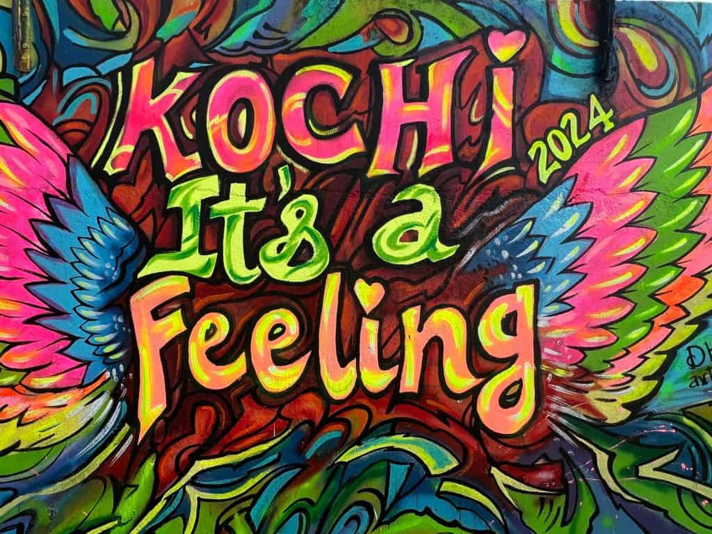 48 hours in Fort Kochi, Bright street art reading 'Kochi it's a feeling' on a wooden board by the beach in Kochi. The colours of paint used are bright green, pink, yellow and blue