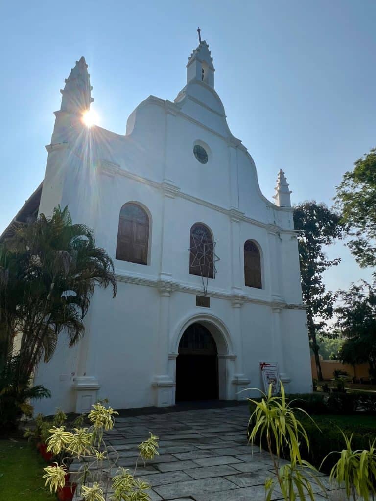 48 hours in Fort Kochi. The white exterior of St Francis Church with the bright blue sky and sun peeking in from behind it. There are green palm trees surrounding the garden area outside.
