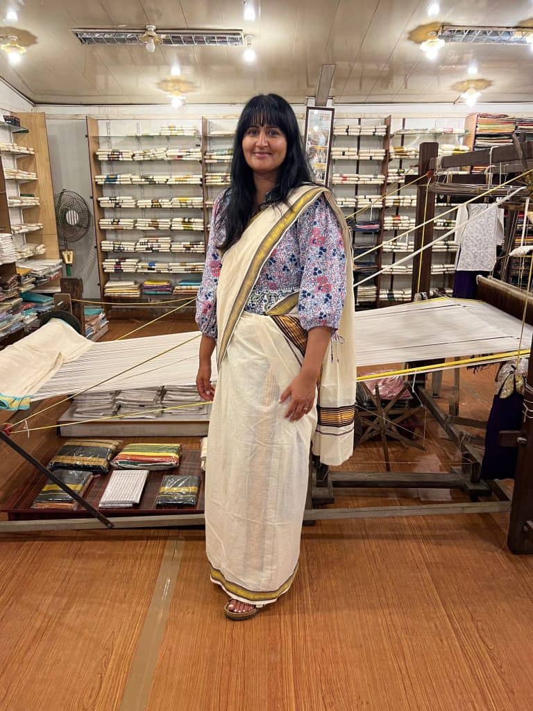 Bejal wearing a traditional white saree in Mattencherry, Fort Kochi. A lady in the background is weaving a saree the traditional way .