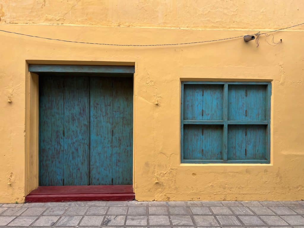 A yellow washed wall with a teal framed window and teal wooden floor. There is a cable running across the top of the exterior. The floor in front of the house is laid with grey paving stones.