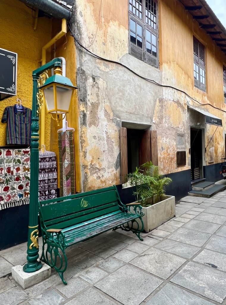 A street in Jew Town with distessed painted yellow and grey walls. There is a green street lamp with is bottle green with yellow accents with a green street bench next to it. Beside the bench is a grey box filled with plants.