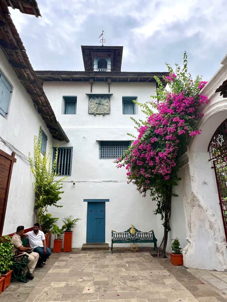 The white washed exterior of the Paradesi Synagoge with Bouganvillea outside. there are 2 men chatting to th eleft on a bench.