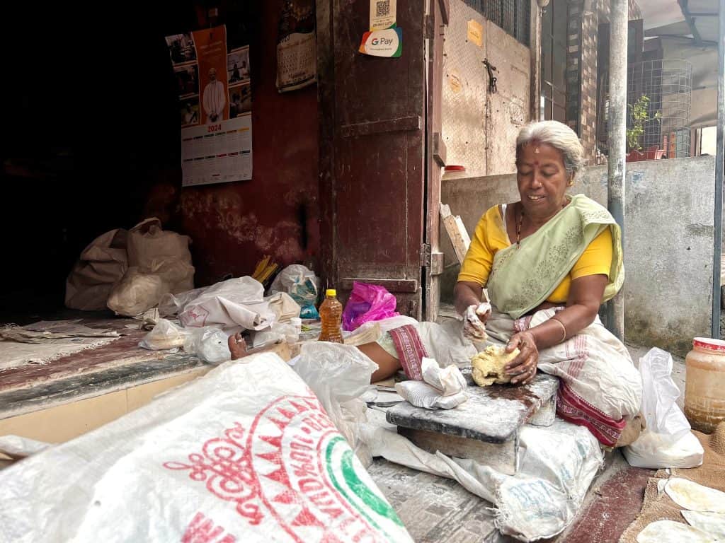 Lady in Fort Kochi rolling out poppadoms outside her shop on the floor. She is wearing a pale green saree and yellow blouse.