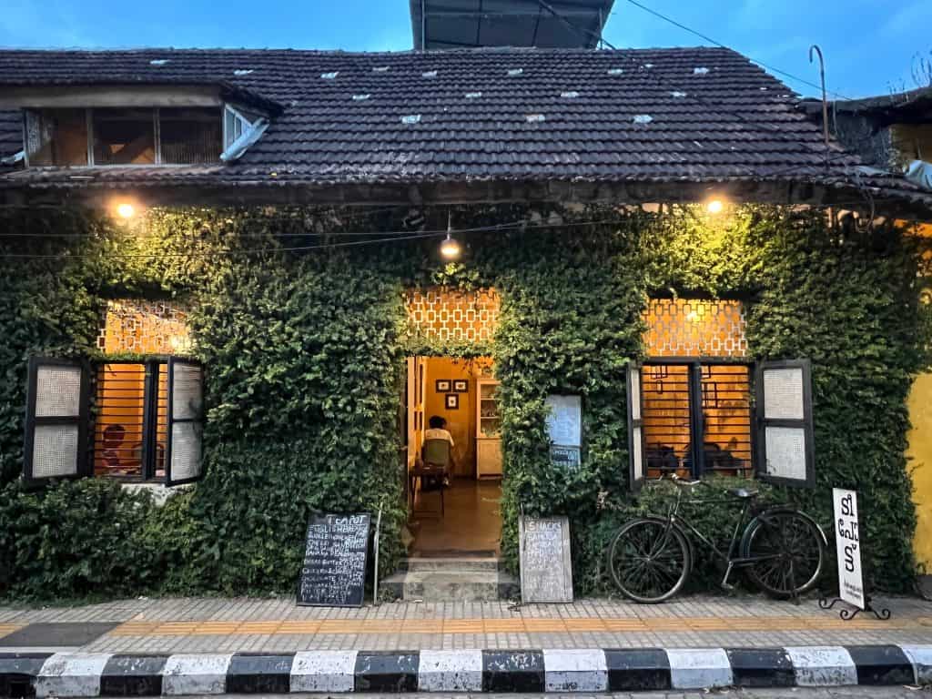 The foliage covered exterior of Teapot Cafe. The lights are on inside and there re people enjoying food and drinks. There are two large plants at either side of the door