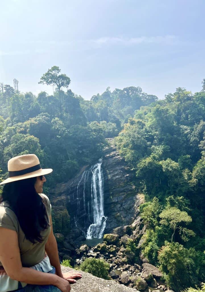 Things to do in Munnar, Valara waterfalls amongst the dense green jungle on the Madurai road from Kochi to Munnar bejal is sitting on the wall to the side of the waterfall wearing a light tan hat and khakhi coloured t-shirt.