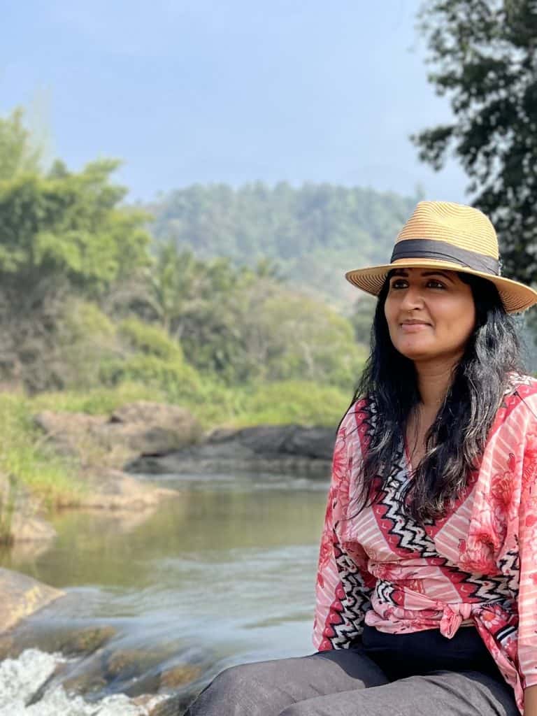 Bejal sitting on a boulder at Ripple waterfalls with the river running behind in Munnar hills. Bejal is wearing a red and pink wrap-over top and tan hat with black ribbon