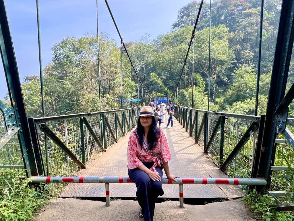 Things to do in Munnar: Bejal sitting at the entrance to the Mattupetty Dam Bridge wearing a tan hat, red and white wrap top and black trousers.