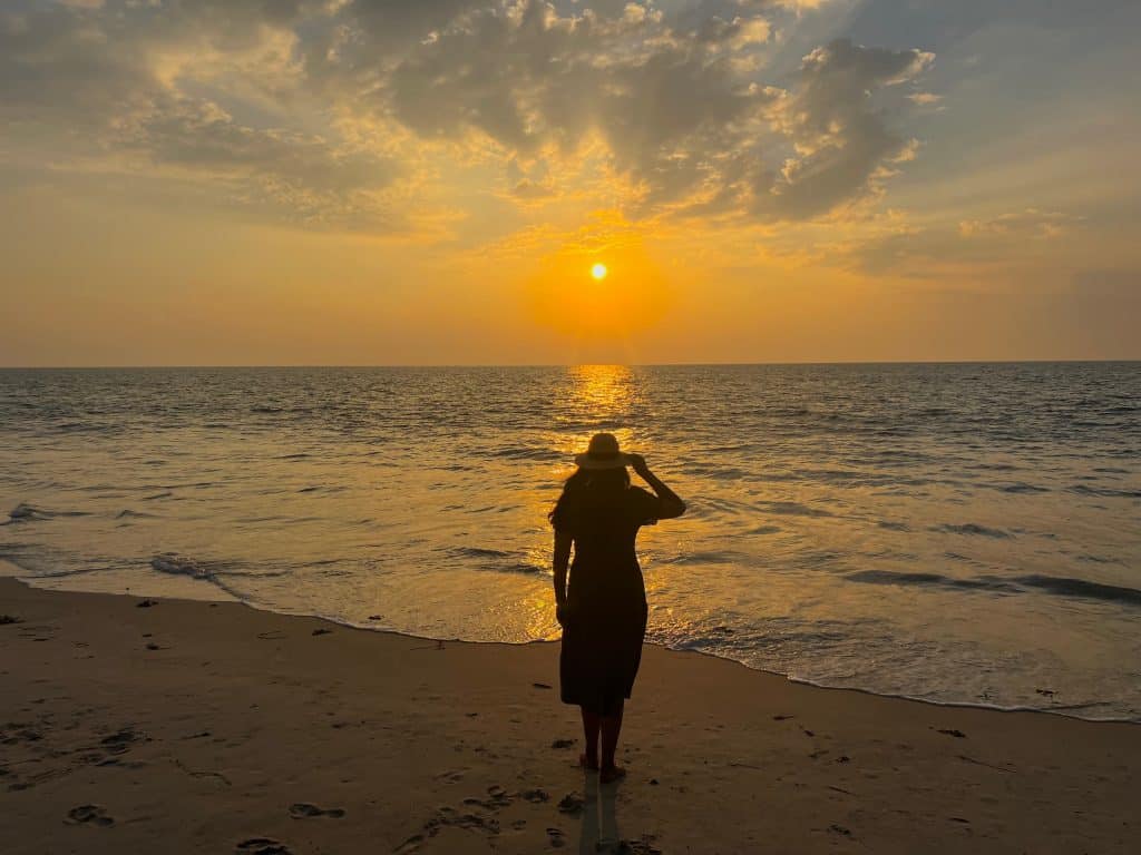 Bejal standing on the sand on Marari beach at sunset with sea lapping against feet. The sun has just about disappeared into the sea line. Grey clouds are in teh sky as the darkness comes