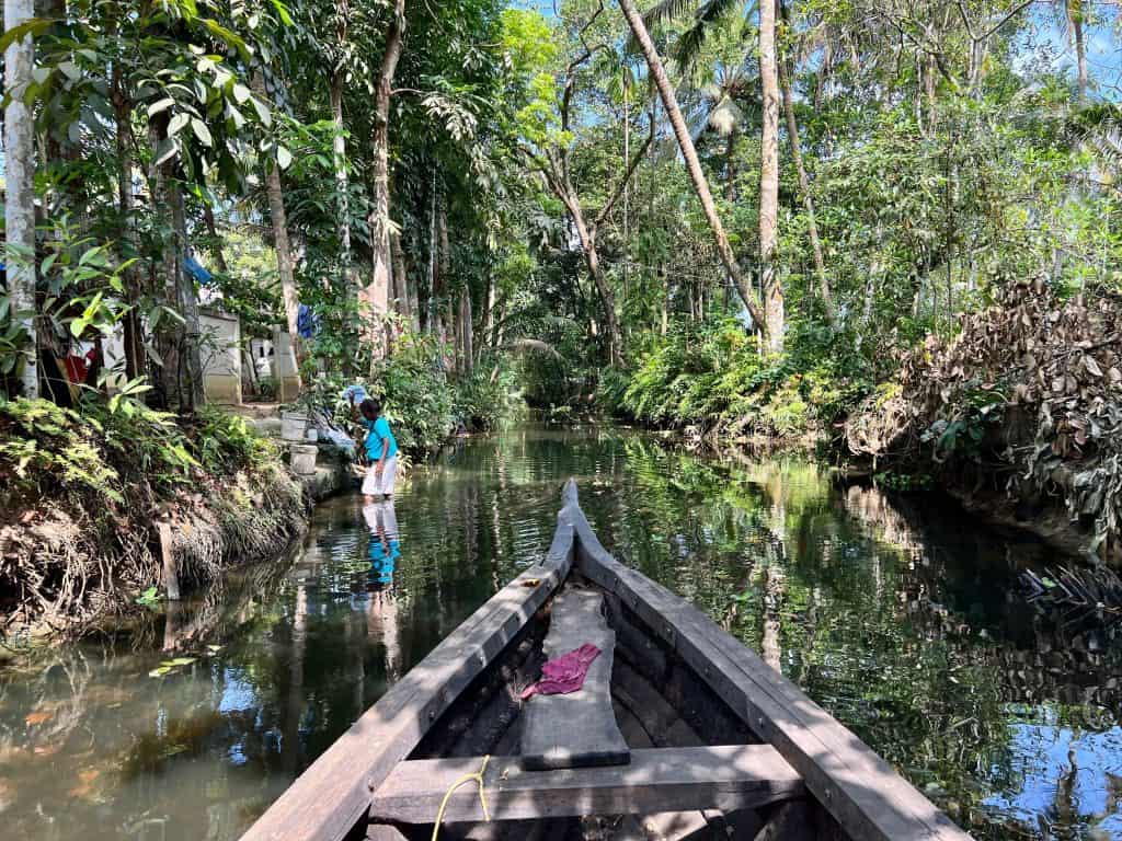 Small canoe-style fishing boat gliding down small canals at Back 2 Village. A lady is doing her washing to the left hand side wearing a blue top. The banks are covered in long palm tree trunks and the lush green foliage of the surrounding forests