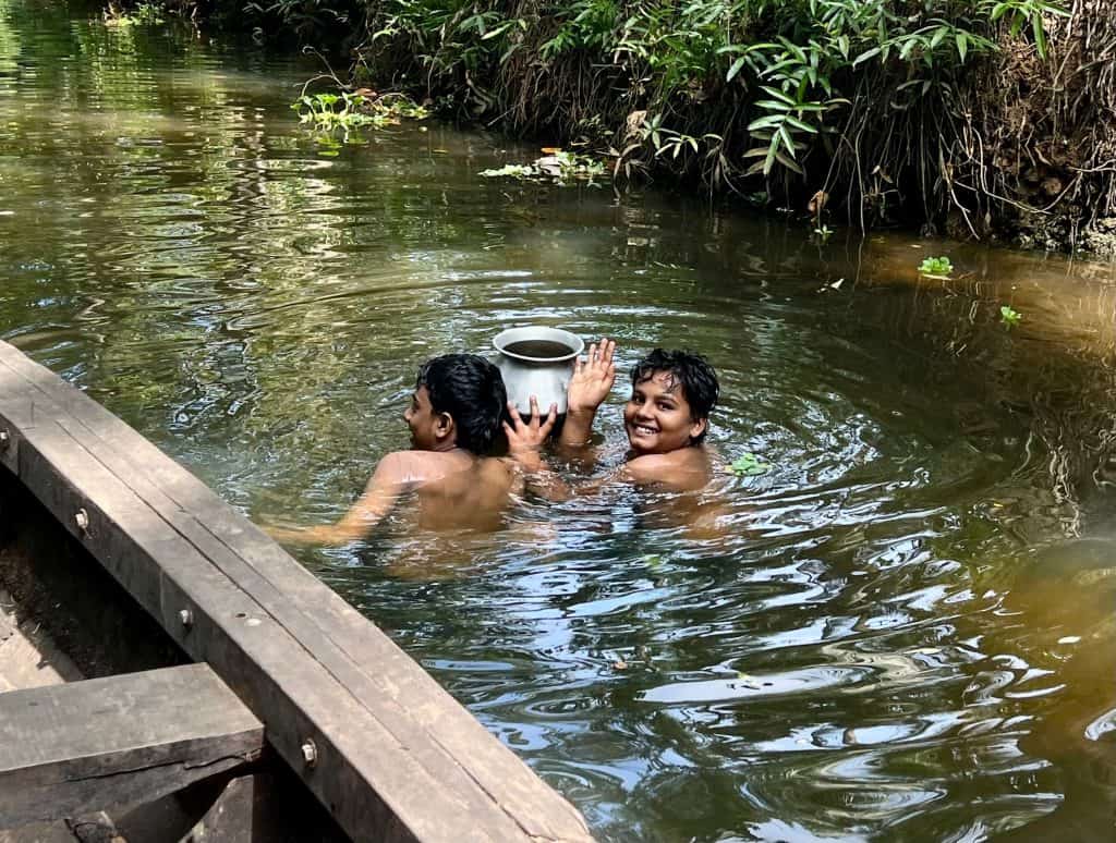 Boys swimming in small canals around Back 2 Village. They are bathing in the river using a steel container to pour wtaer on themselves. Our canoe passes by one side of them and the river bank with foliage is on the right hand side