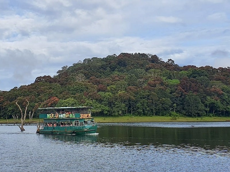 A green boat with 2 levels filled with tourists at Periyar National Park in Kerala. The boat is floating along the lake and there are trees and mountains in the background
