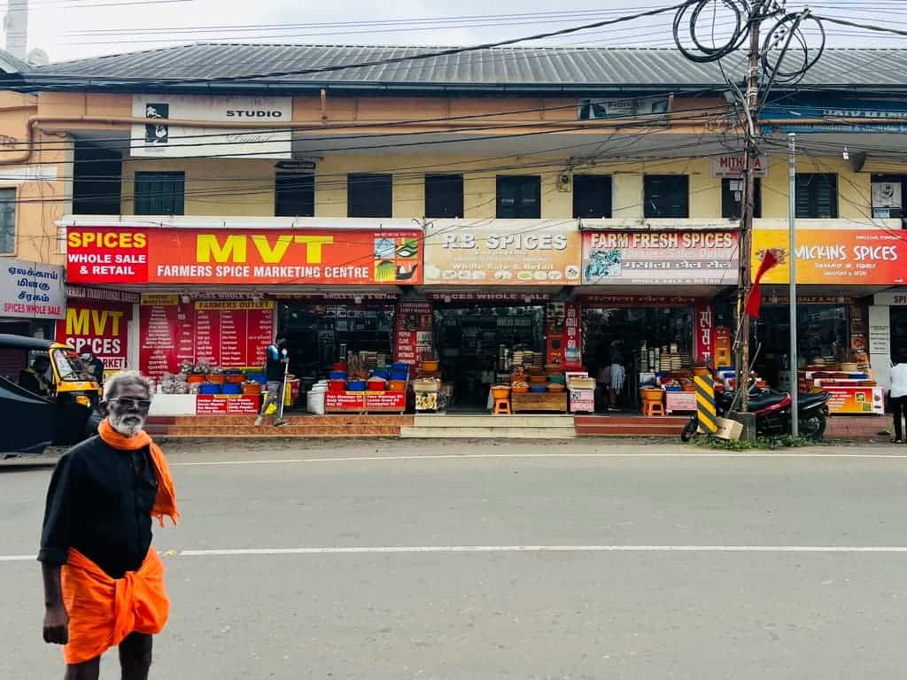 Man wearing an orange material around his lower half standing infront of a row of shops with 'MVT' on the board in the centre of Thekkady, Kerala