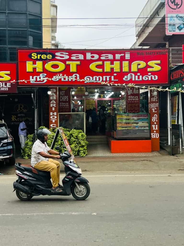 A man on a scooter riding past a red shop sign the Thekkady centre, Kerala, reading 'Hot Chips' in yellow.